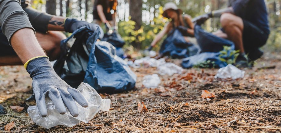 Close up of hands with plastic bottle with group of young people friends volunteers collecting plastic bottles to trash bags in forest background. Ecology concept